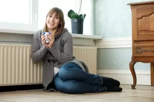 Woman holding mug and sitting on floor, leaning against heater along wall. 