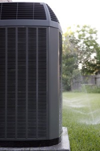 Close-up of a black HVAC unit on a slab outside a home. Green grass surrounding and sprinklers turned on in background.