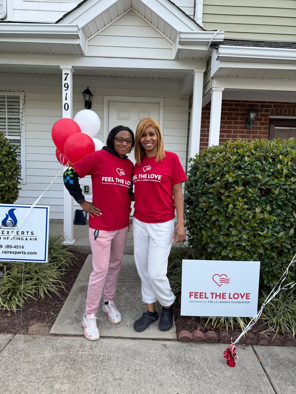 Two smiling Feel the Love volunteers in red Feel the Love shirts stand in front of a home with red and white balloons, an Air Experts yard sign and a Feel the Love yard sign