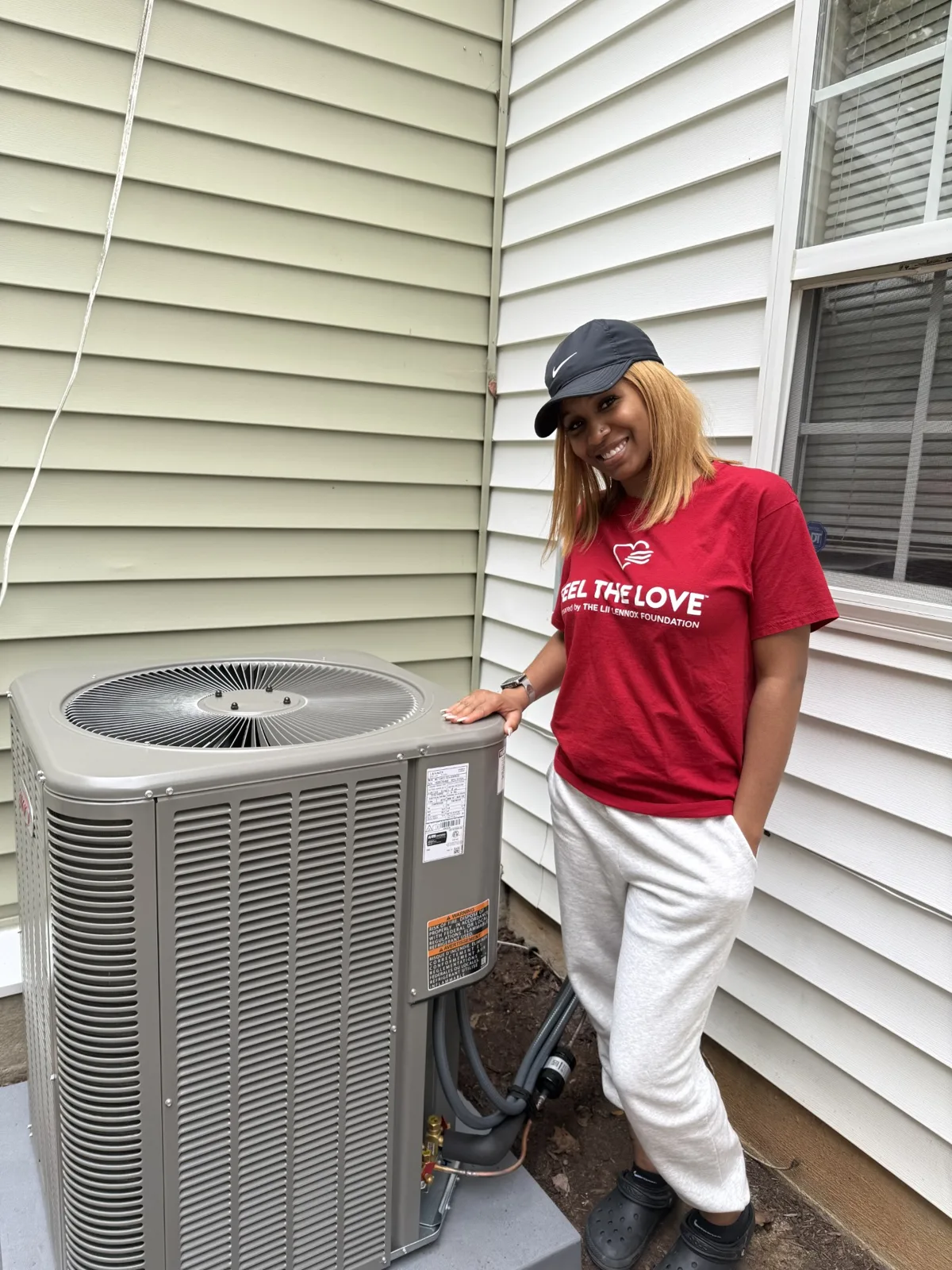 Smiling Feel the Love volunteer in a red Feel the Love shirt standing beside the newly installed Lennox outdoor unit