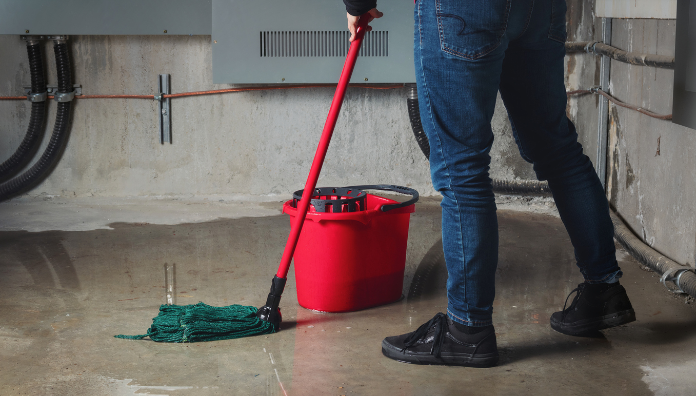 Water damage from rain, snowmelt or pipe burst coming from multiple cracks and leaks in concrete wall and ceiling. Person mopping up water with red mop and bucket.