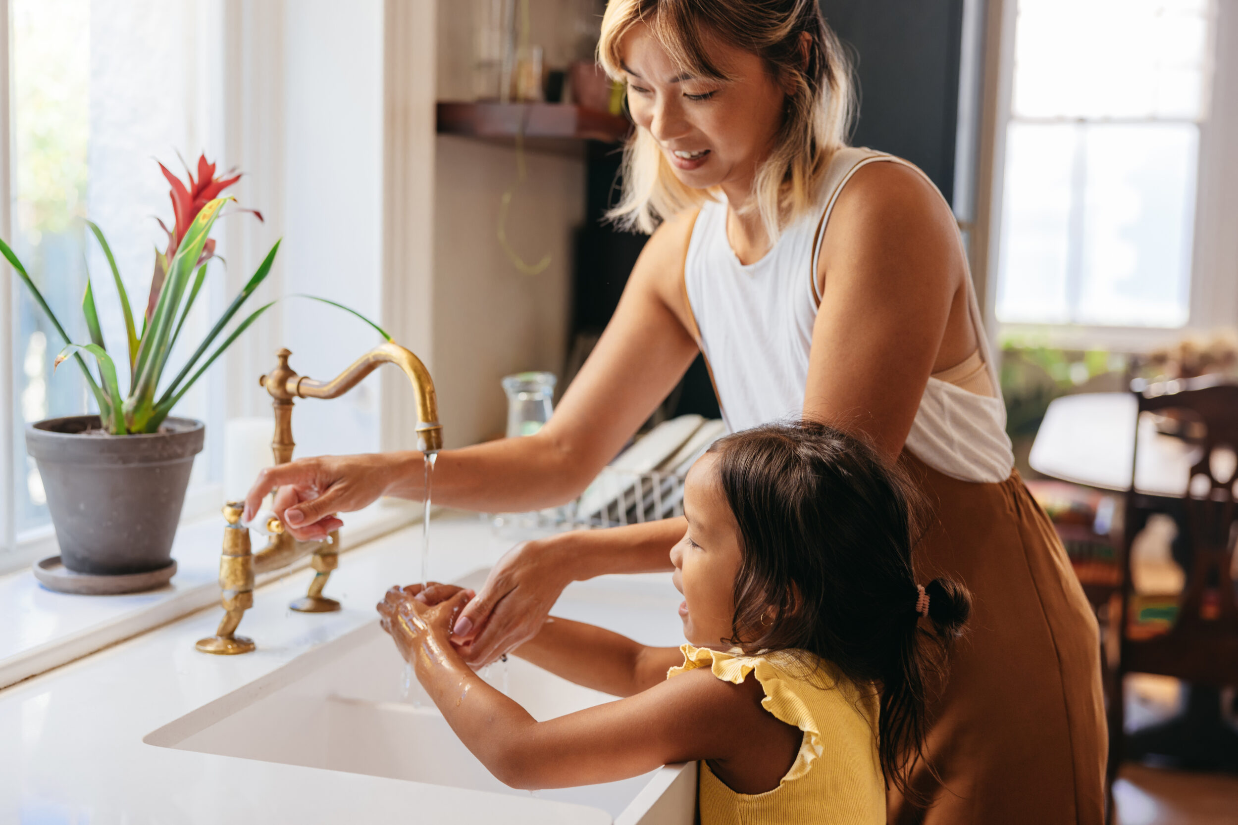 Young woman teaching her daughter to wash her hands with soap at kitchen sink. White countertop, gold faucet.